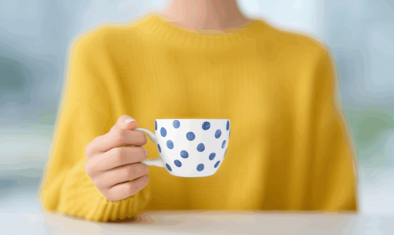 Keltainen villapaita naisen yllä ja valkoinen kahvikuppi sinisillä koristeilla käsissä – rauhallinen hetki, joka kuvastaa graafisen suunnittelun ja visuaalisen viestinnän lämpöä ja yhtenäisyyttä.