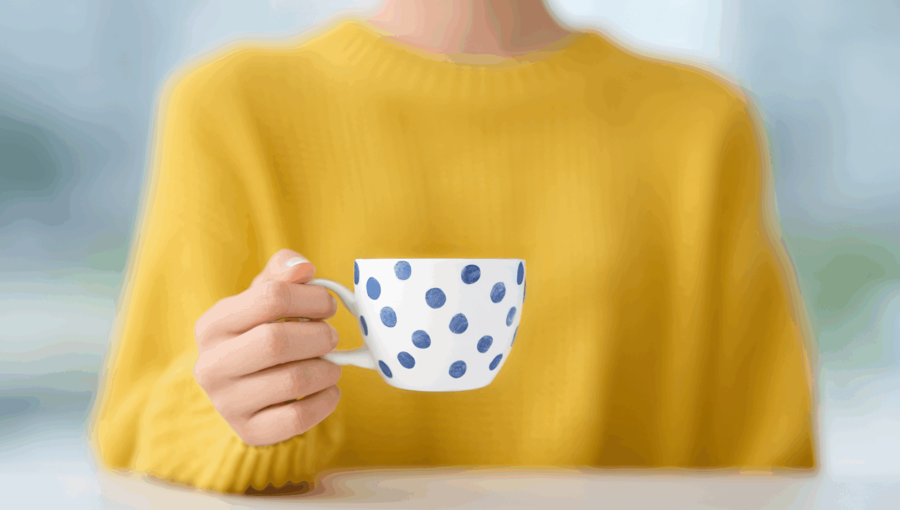 Keltainen villapaita naisen yllä ja valkoinen kahvikuppi sinisillä koristeilla käsissä – rauhallinen hetki, joka kuvastaa graafisen suunnittelun ja visuaalisen viestinnän lämpöä ja yhtenäisyyttä.